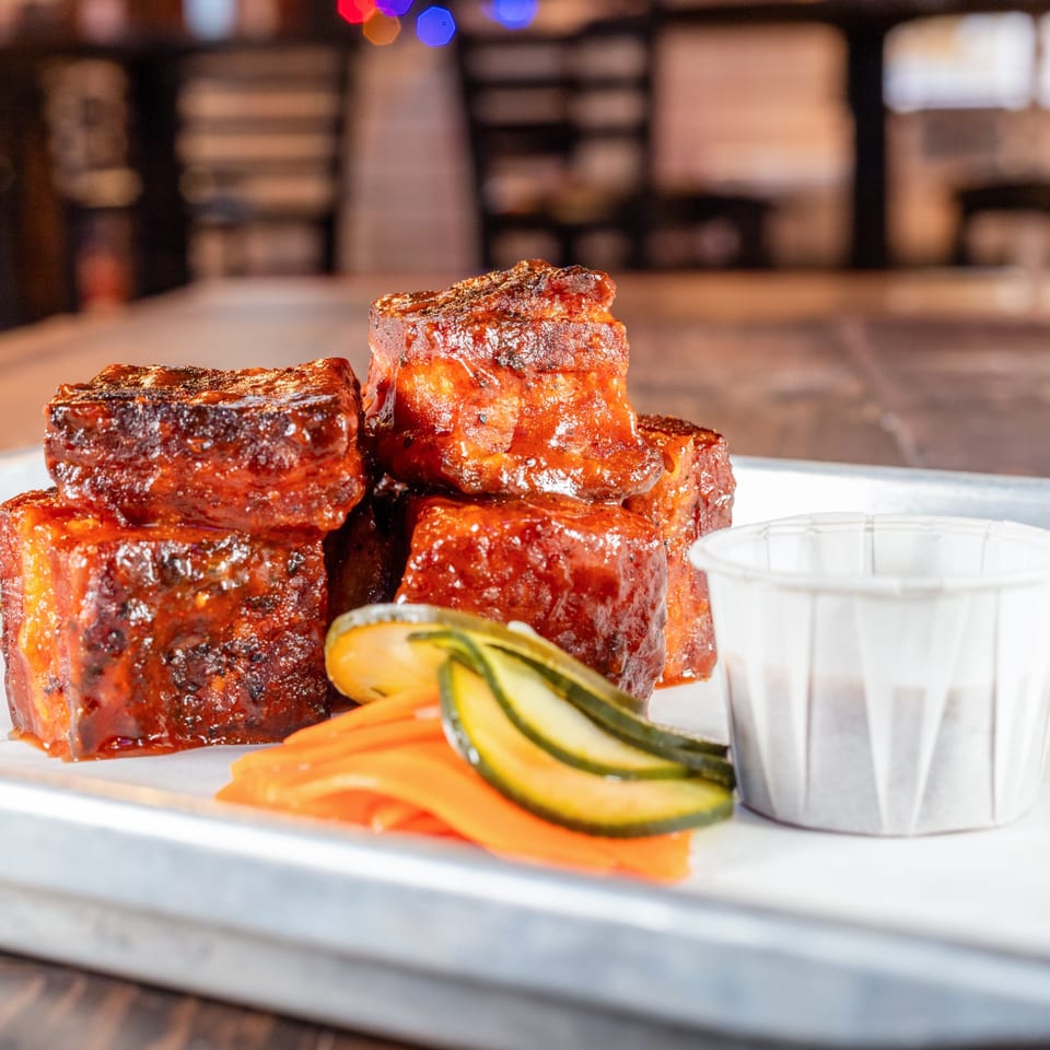 Hattie's BBQ burnt ends, glazed in sauce, with pickled carrots and cucumbers on a tray.